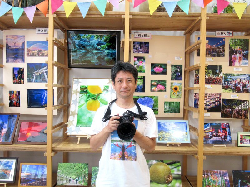 夏を感じさせる写真が並ぶ作品展と白井さん（中央）＝市川市