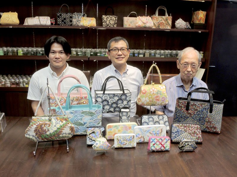 鮮やかな刺しゅうの施されたビーズバッグなどの商品を前に、変わらぬ物作りへの心意気を語る仙田社長（中央）と先代の秀一さん（右）、次代を担う晃さん＝柏市