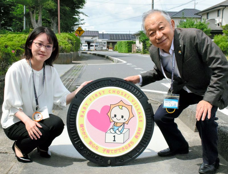 赤ちゃん太陽くんが愛らしい新型マンホールふたを紹介する小高村長（右）と鈴木さん＝長生村