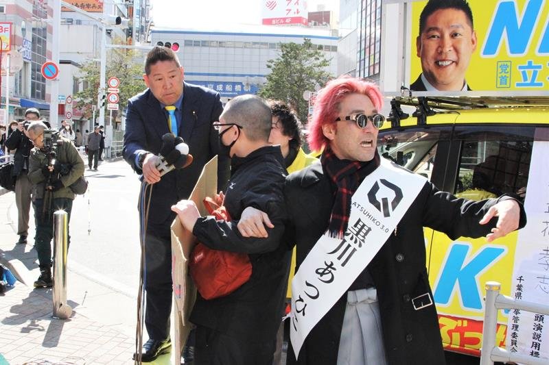 千葉県知事選の第一声で、抗議のプラカードを掲げる聴衆にマイクを向ける立花氏（左）と、聴衆を誘導する黒川氏（右）＝２月２７日、ＪＲ船橋駅前
